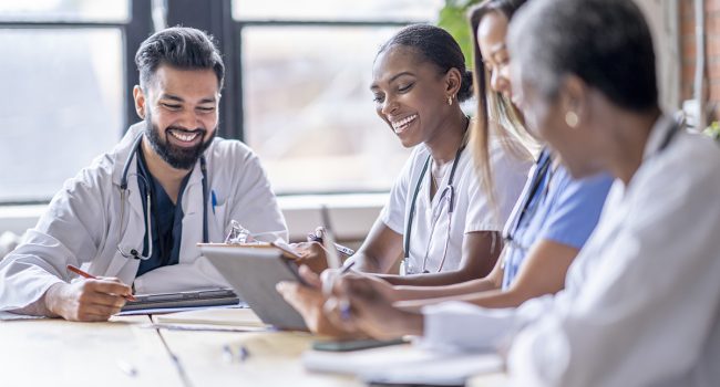 A small group of four medical professionals sit around a boardroom table as they meet to discuss patient cases.  They are each dressed professionally in scrubs and lab coats as they focus on working together.